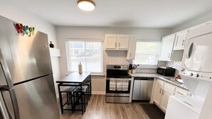 A compact kitchen with a refrigerator, stove, microwave, and sink. There is a dining table with chairs and a window with blinds.
