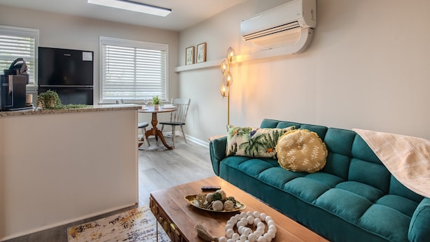 Ein modernes Wohnzimmer mit einem türkisfarbenen Sofa, einem Holztisch und einer Küchenzeile mit Kühlschrank und Fernseher.