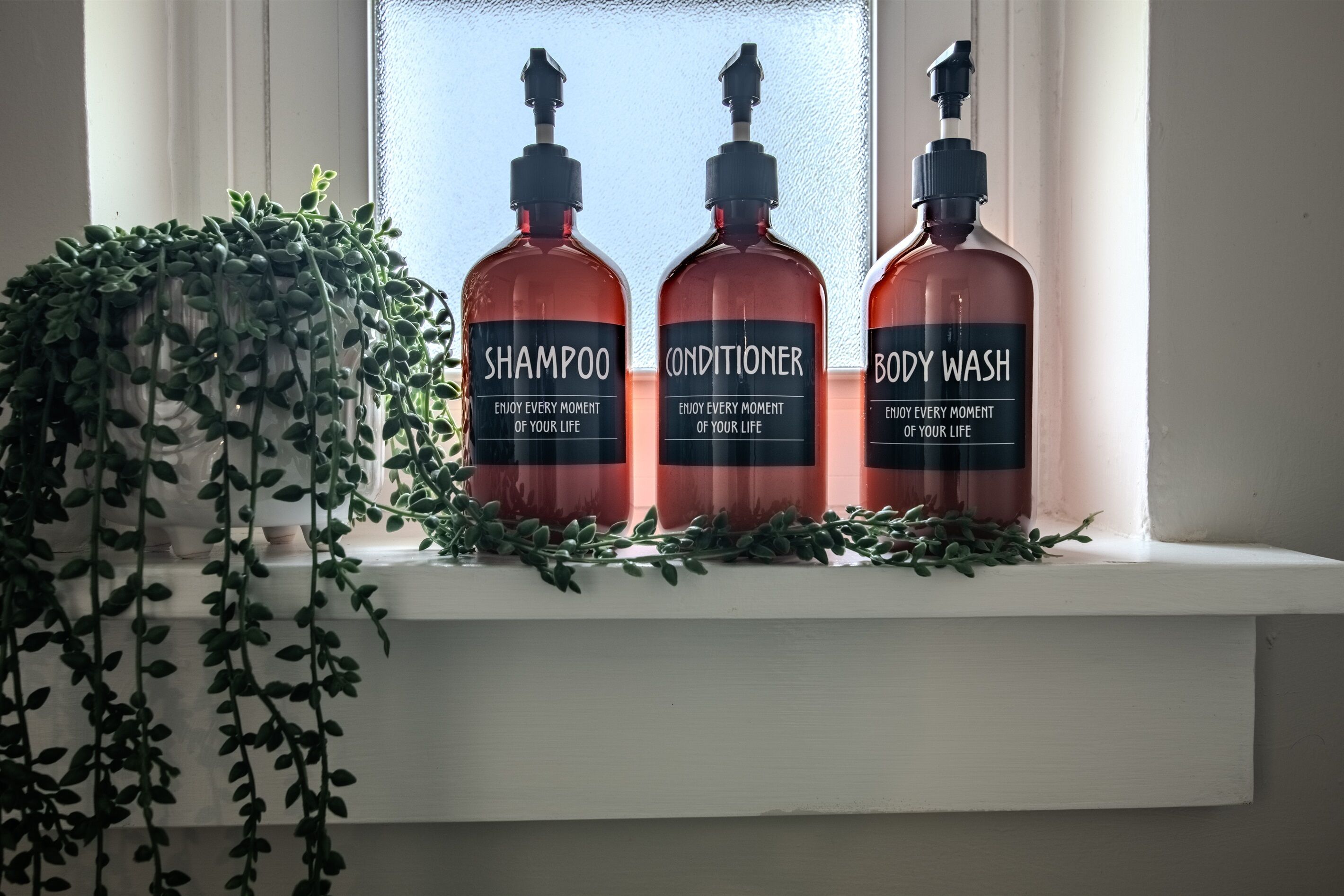 Trois bouteilles de shampoing, d’après-shampoing et de gel douche sont posées sur un rebord de fenêtre, à côté d’une plante.