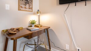Un espacio de trabajo moderno con un escritorio de madera, una silla de vidrio transparente, una lámpara, una planta en maceta y un televisor montado en la pared.