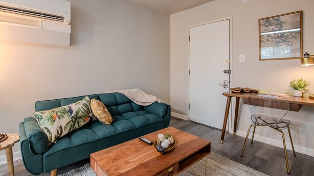 Una sala de estar con un sofá verde, una mesa de centro de madera, una mesita auxiliar y un aire acondicionado instalado en la pared.