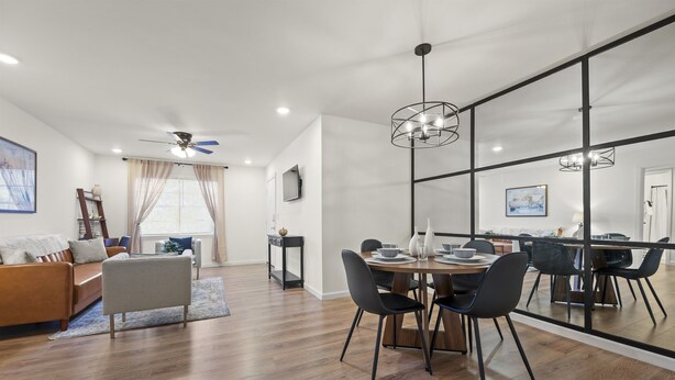 Ein modernes Wohnzimmer mit Sofa, Esstisch mit Stühlen, Deckenventilator und großem Fenster mit Vorhängen.