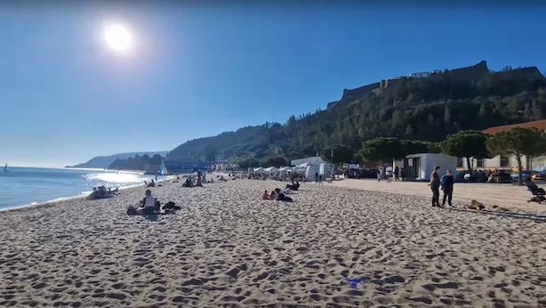 A beach with people sunbathing and walking, a clear sky, and a distant hill.