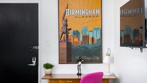A room with a black door, a poster of Birmingham, Alabama, a desk with a lamp, a pink chair, and a small plant.