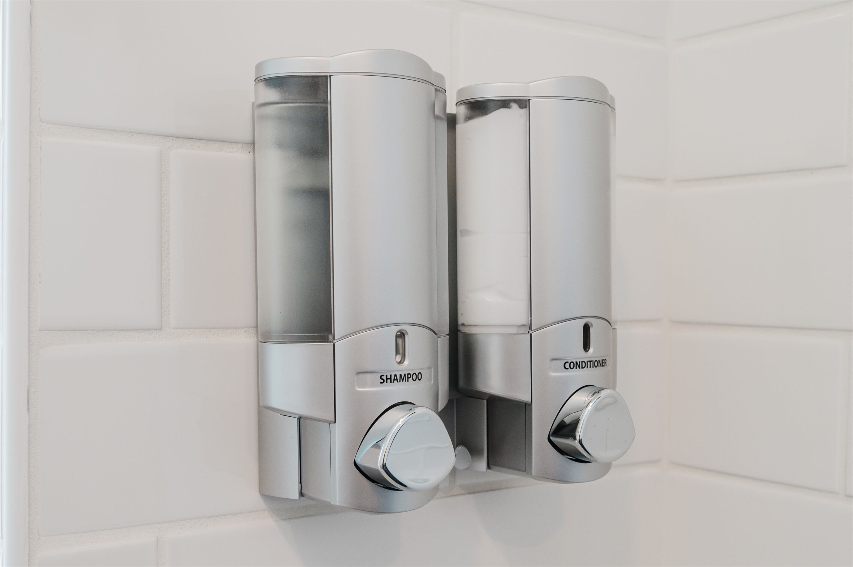 Zwei an der Wand befestigte Spender, einer für Shampoo und einer für Conditioner, in einem Badezimmer mit weißen Fliesen.