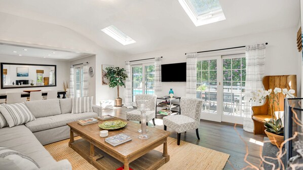 A spacious living room with a sofa, coffee table, and chairs, large windows with curtains, and a flat-screen TV.