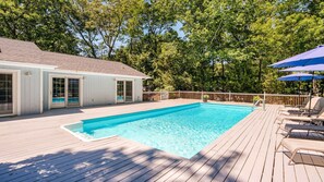 Une piscine avec une terrasse en bois, un patio couvert avec des chaises et une maison dotée de portes-fenêtres coulissantes.