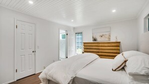 A bedroom with a bed, a wooden headboard, a white door, and a window with a beach view.