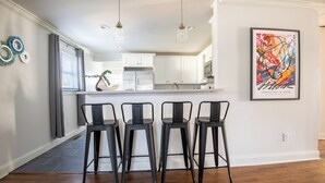 A modern kitchen with white cabinets, a central island, and three black bar stools. - Crestwood Bloom – 3BR Getaway w/ Patio & Backyard (birmingham)