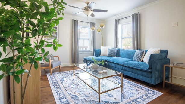 A living room with a blue sofa, a glass coffee table, a ceiling fan, and a large window with curtains. - Crestwood Bloom – 3BR Getaway w/ Patio & Backyard (birmingham)