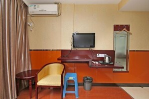 A compact hotel room with a single bed, a desk with a computer, a chair, a small stool, a trash bin, and a television mounted on the wall.