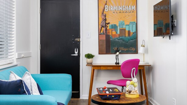 A room with a black door, a blue sofa, a pink chair, a small table with decorative items, and two wall-mounted posters. - D3-Vulcan's Double Suite-King Beds/Workspace/Pets (Birmingham)