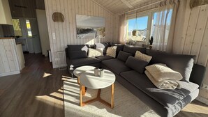 A cozy living room with a dark grey sectional sofa, a round coffee table, and a large window with curtains. - North Sea vacation home Huus Eiderente - relaxation, wellness, fireplace, fiber optic WLAN (Wesselburenerkoog)