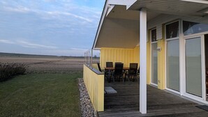 A covered outdoor area with a wooden deck, a yellow wall, and a view of a field. - North Sea vacation home Huus Eiderente - relaxation, wellness, fireplace, fiber optic WLAN (Wesselburenerkoog)