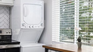 A compact kitchen with a built-in oven, a microwave, and a washing machine. There is a window with blinds, a small table, and a bench.