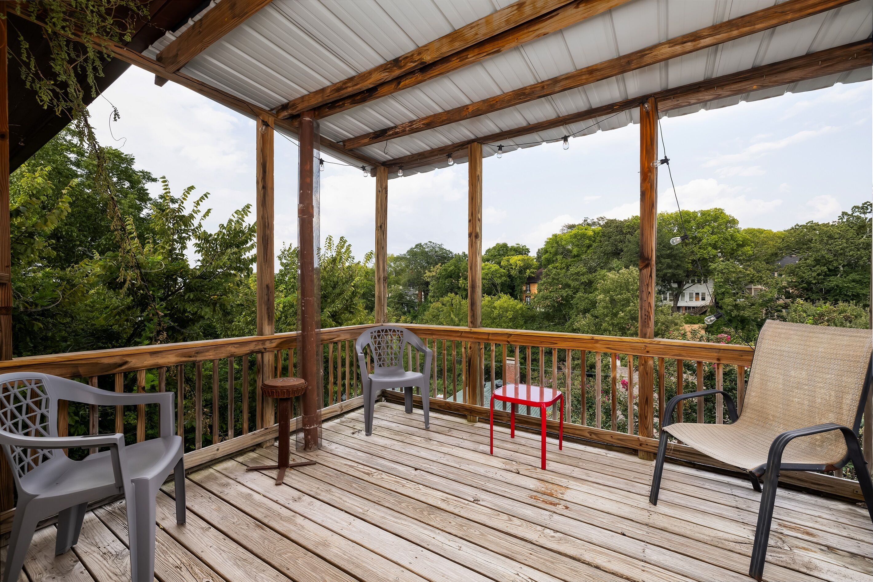 A wooden deck with a metal roof, surrounded by greenery and a distant house.