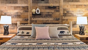 A bedroom with a wooden wall, a bed with a patterned bedspread, two bedside tables with lamps, and a shelf with decorative items.