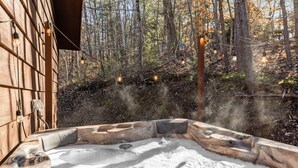 A hot tub with a wooden exterior, surrounded by a forested area with string lights.