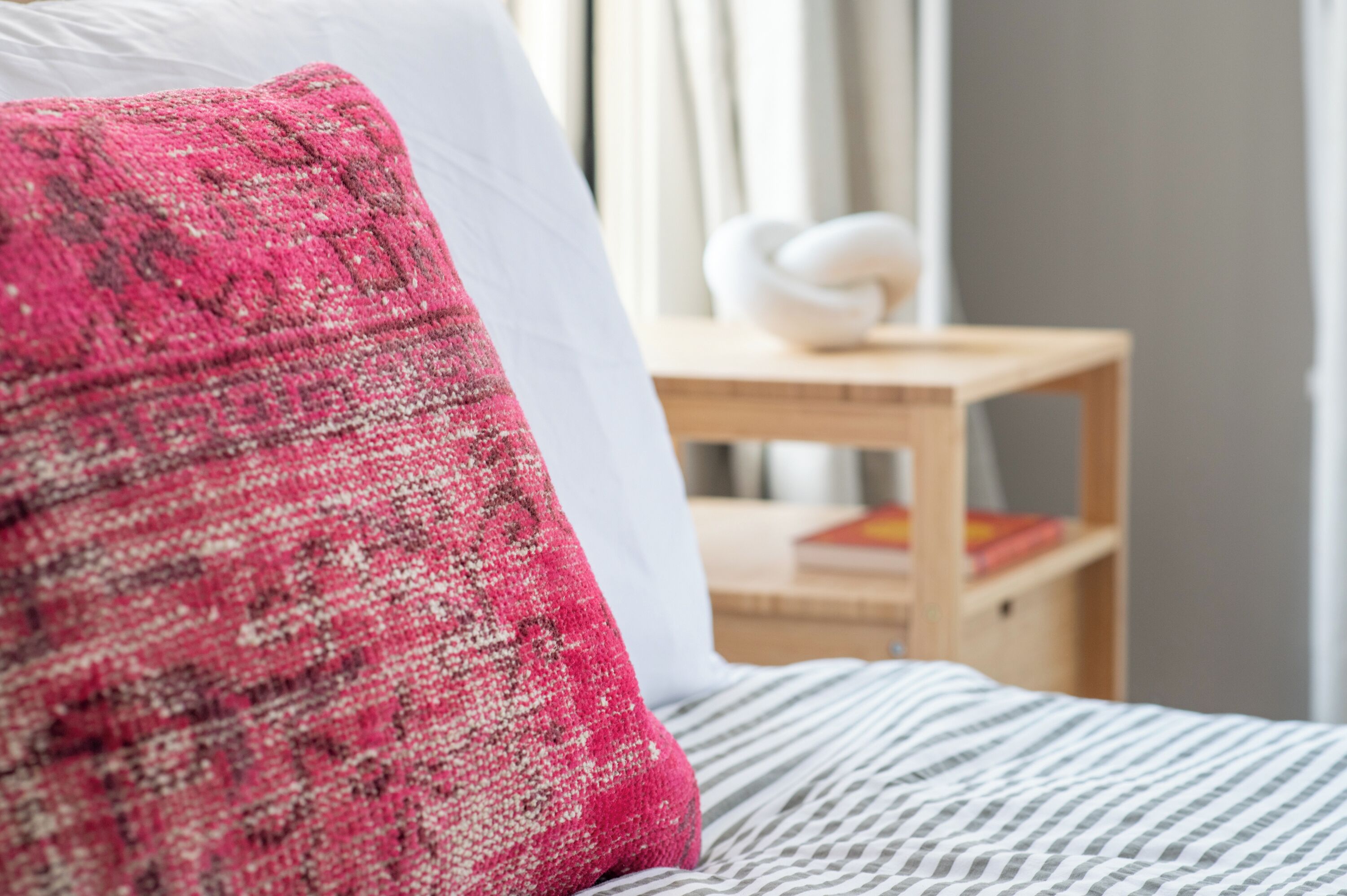 Un lit avec un oreiller à motifs roses et blancs, un oreiller blanc et une couvre-lit à rayures. On aperçoit une table de chevet en bois, sur laquelle se trouve un objet décoratif blanc, ainsi que des livres en arrière-plan.