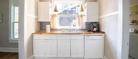 Cocina compacta con armarios blancos, encimera de madera, cafetera y ventana con cortinas.