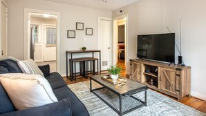 A living room with a dark-colored sofa, a coffee table with a plant, a wooden TV stand, and a ceiling fan. - Vulcan View- Groups/King Beds/Free Parking/2 Units (Birmingham)