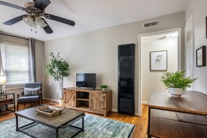 Un salon avec un ventilateur de plafond, un téléviseur posé sur un meuble en bois, un fauteuil, une table basse et une plante en pot.