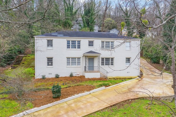 Une maison à deux étages avec un porche à l’avant, une allée et une cour avec des arbres et des arbustes.