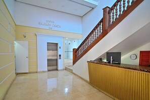 A hotel lobby with a reception desk, a staircase, a clock, and a door labeled 'EDIFICIO SALOMON GANEM 1948'.