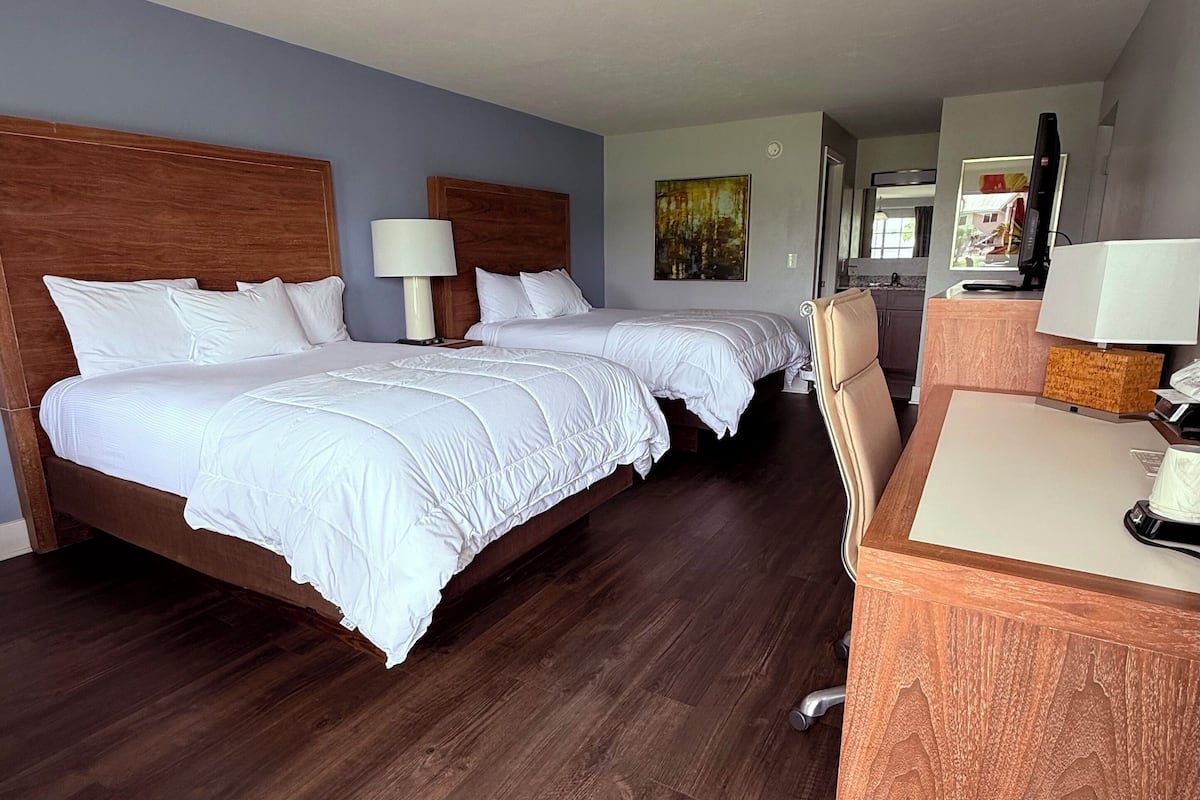 Habitación de hotel con dos camas, un escritorio, una silla, una lámpara y un cuadro en la pared.