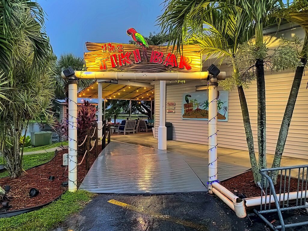 A tiki bar entrance with a neon sign, palm trees, and a covered patio area.