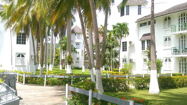 A white multi-story hotel with balconies and a well-maintained garden with palm trees and flowers.