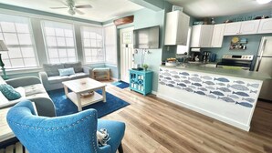A living room with a blue armchair, a sofa, a coffee table, and a TV. The kitchen has white cabinets, a fridge, and a stove. - Cozy Cottage At Lake Weir (Ocklawaha)