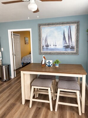 A dining area with a wooden table, two grey cushioned stools, a ceiling fan, a framed painting of sailboats, a potted plant, and a trash can.