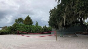 Un terrain de volleyball de plage, avec du sable et un filet rouge, entouré d’arbres et avec un bâtiment visible au loin.