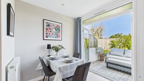 A dining area with a table set for two, a framed floral painting on the wall, and a view of the garden through a sliding glass door. - Charming 2-bed Family Cottage Your West Bay Retreat (Yarmouth)