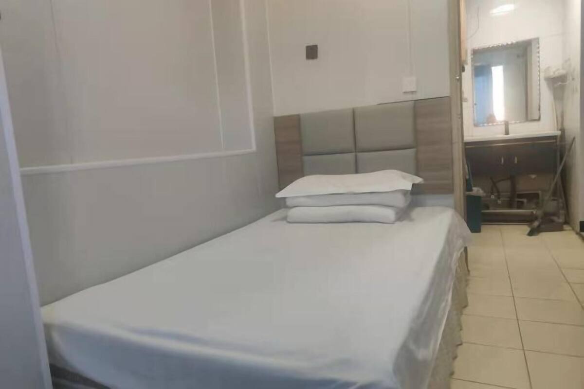 Una habitación pequeña con una cama individual, una colcha blanca sencilla, un cabecero con sección acolchada y una mesita de noche con lámpara.