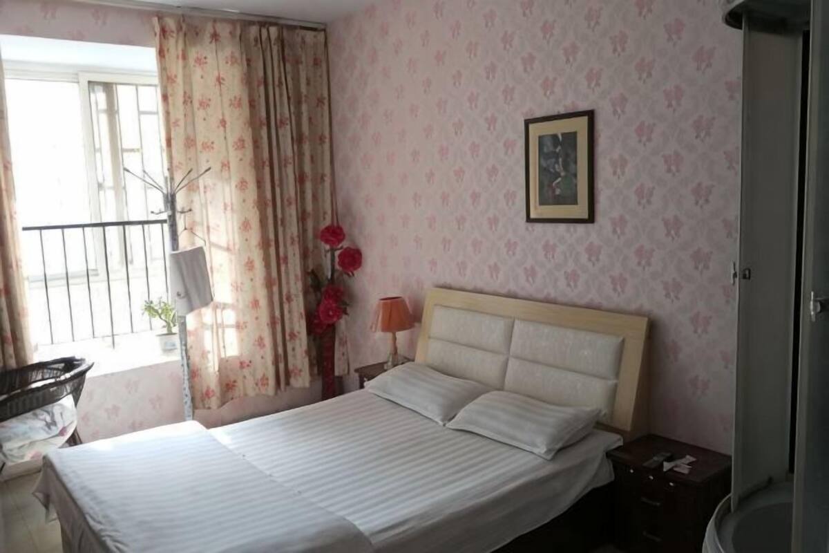 Un dormitorio con cama, mesita de noche, ventana con cortinas, un cuadro enmarcado en la pared y una pequeña mesa con lámpara.
