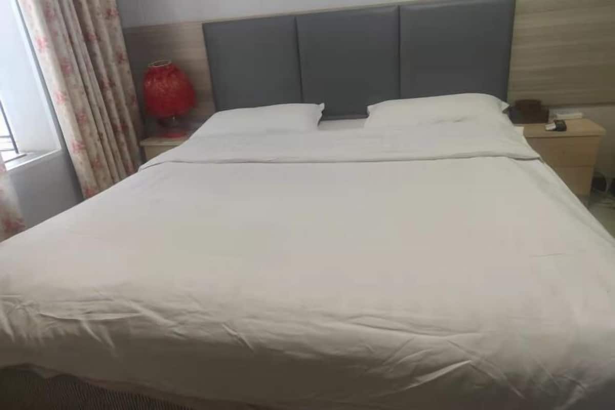 Una cama bien hecha con un cabecero gris, una mesita de noche con una lámpara y una ventana con cortinas.