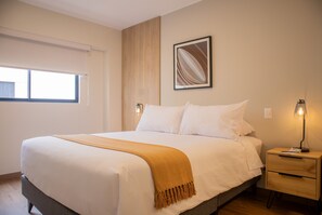 A neatly made bed with a white comforter and a mustard-colored throw blanket. There is a wooden nightstand with a lamp and a small drawer. A framed artwork hangs on the wall above the bed.