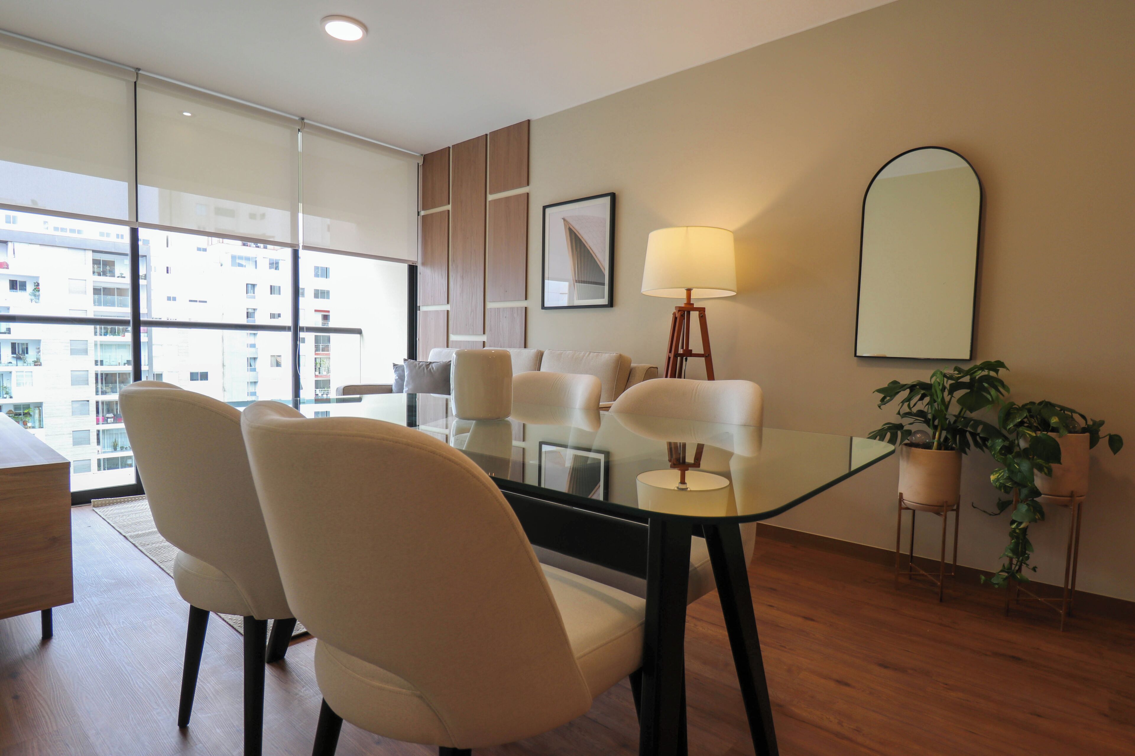 A modern hotel room with a glass dining table, white chairs, a standing lamp, and a large window offering a city view.
