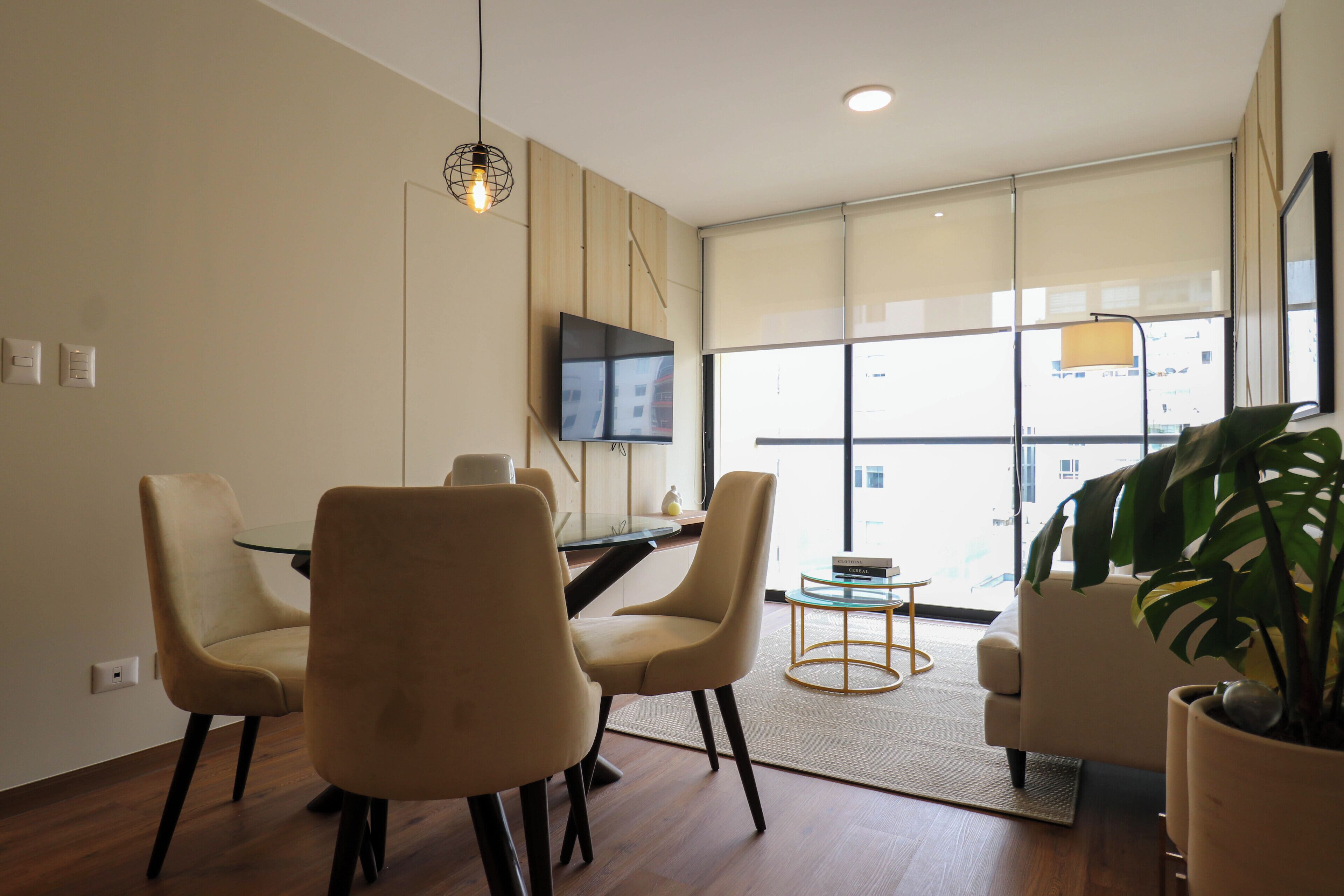 Una sala de estar moderna con comedor, televisor montado en la pared y un ventanal con vista a la ciudad.