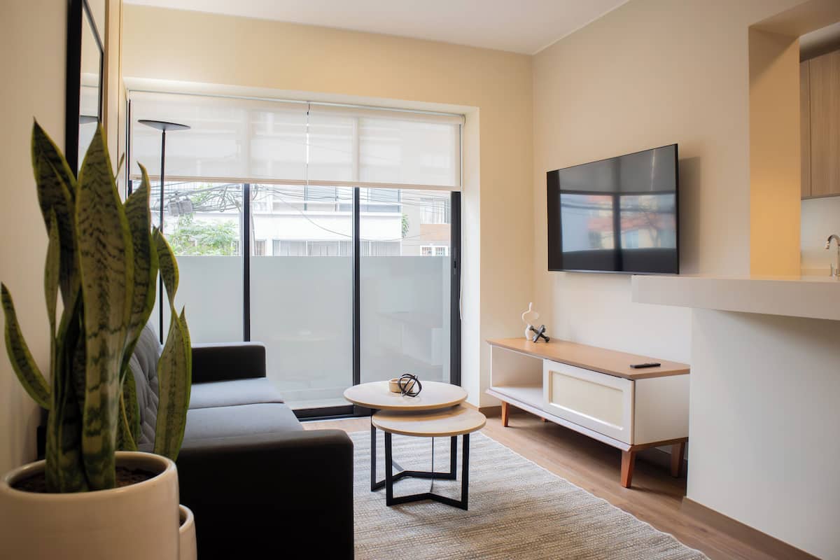 Un salon moderne avec un canapé, une table ronde, une plante en pot, un téléviseur fixé au mur et une porte-fenêtre coulissante donnant sur un balcon.