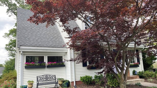 Una casa de dos pisos con techo gris, revestimiento blanco y un porche delantero. Hay macetas con flores en las ventanas, un banco y un árbol con hojas rojas.