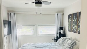 Un dormitorio con cama, ventilador de techo, televisor, cafetera y ventana con persianas.
