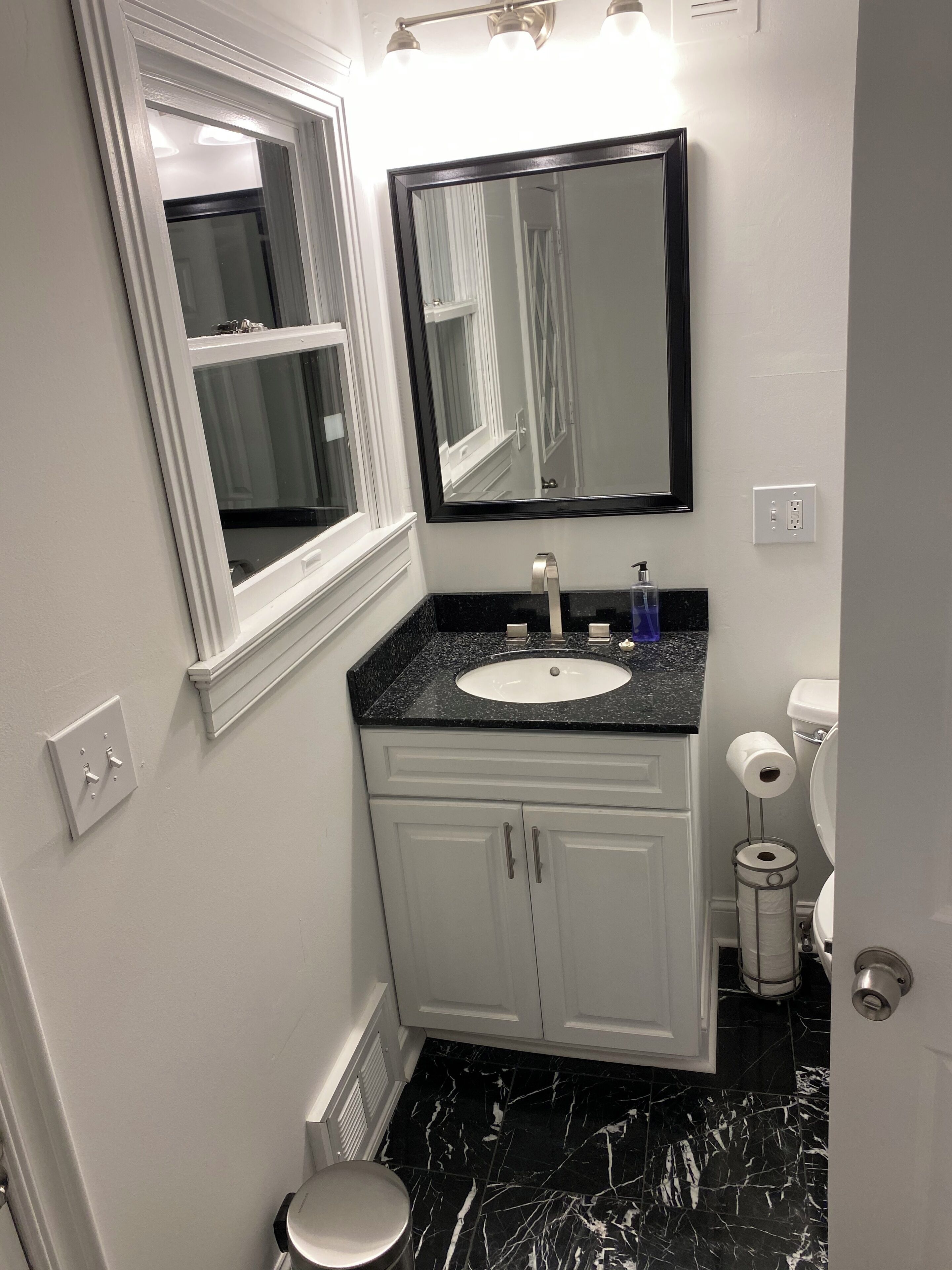 Een badkamer met een wit wastafelmeubel, een vloer van zwart marmer, een grote spiegel en een toilet met een papierhouder.