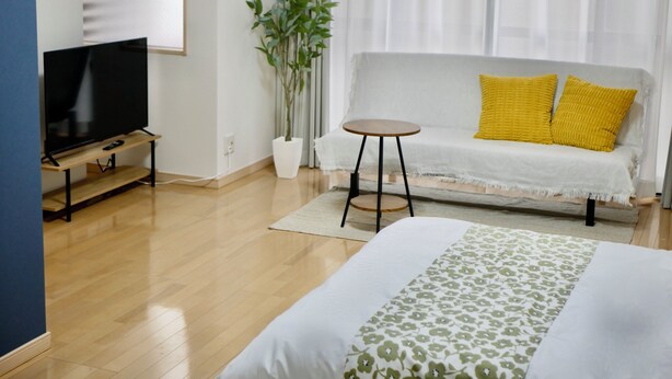 Une chambre d’hôtel moderne avec un téléviseur à écran plat posé sur un meuble en bois, un canapé gris agrémenté de coussins jaunes, une plante en pot et un lit recouvert d’une courtepointe à motifs floraux.