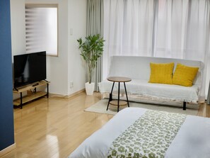 Quarto de hotel moderno com televisão de tela plana sobre suporte de madeira, sofá cinza com almofadas amarelas, planta em vaso e cama com colcha florida.