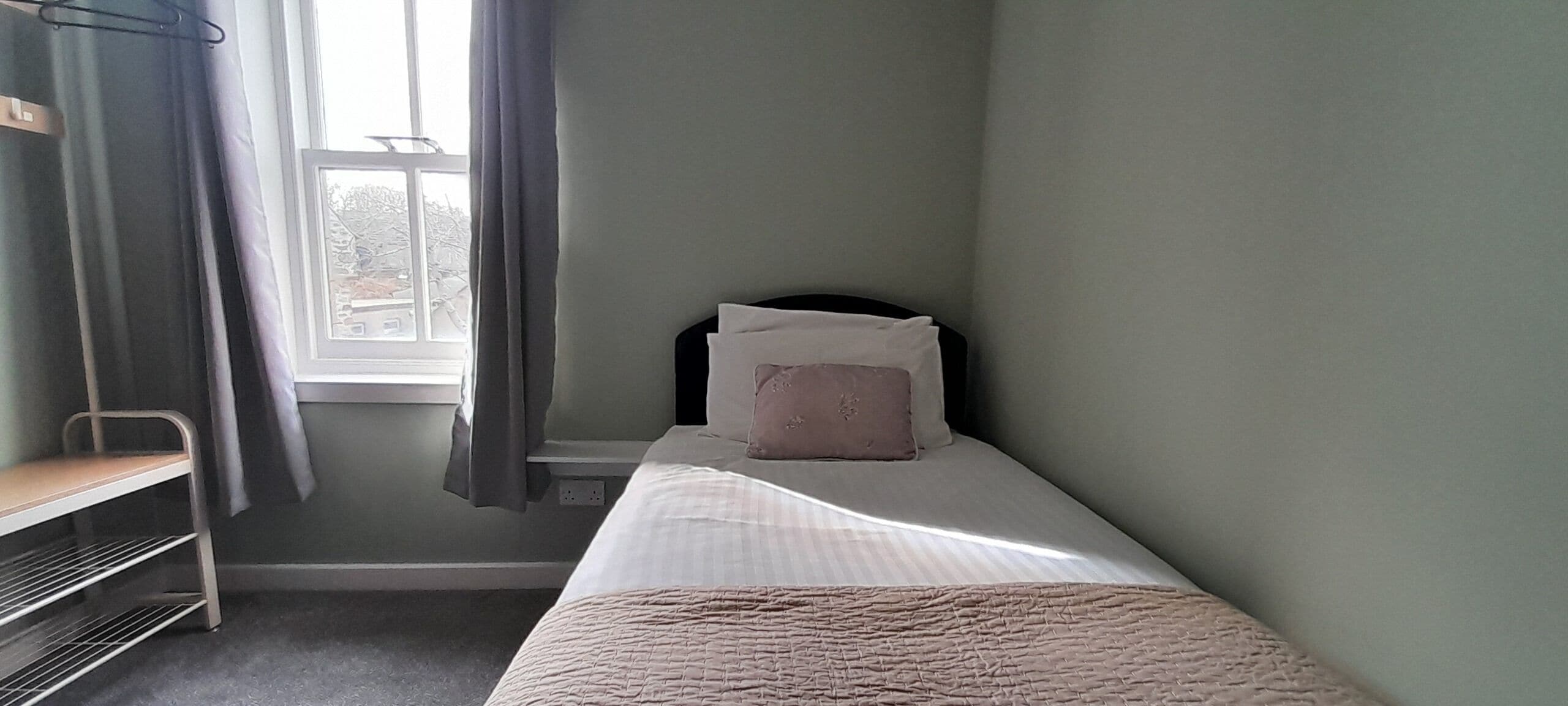 Una habitación pequeña con una cama individual, una ventana, un estante metálico y una pared sencilla.