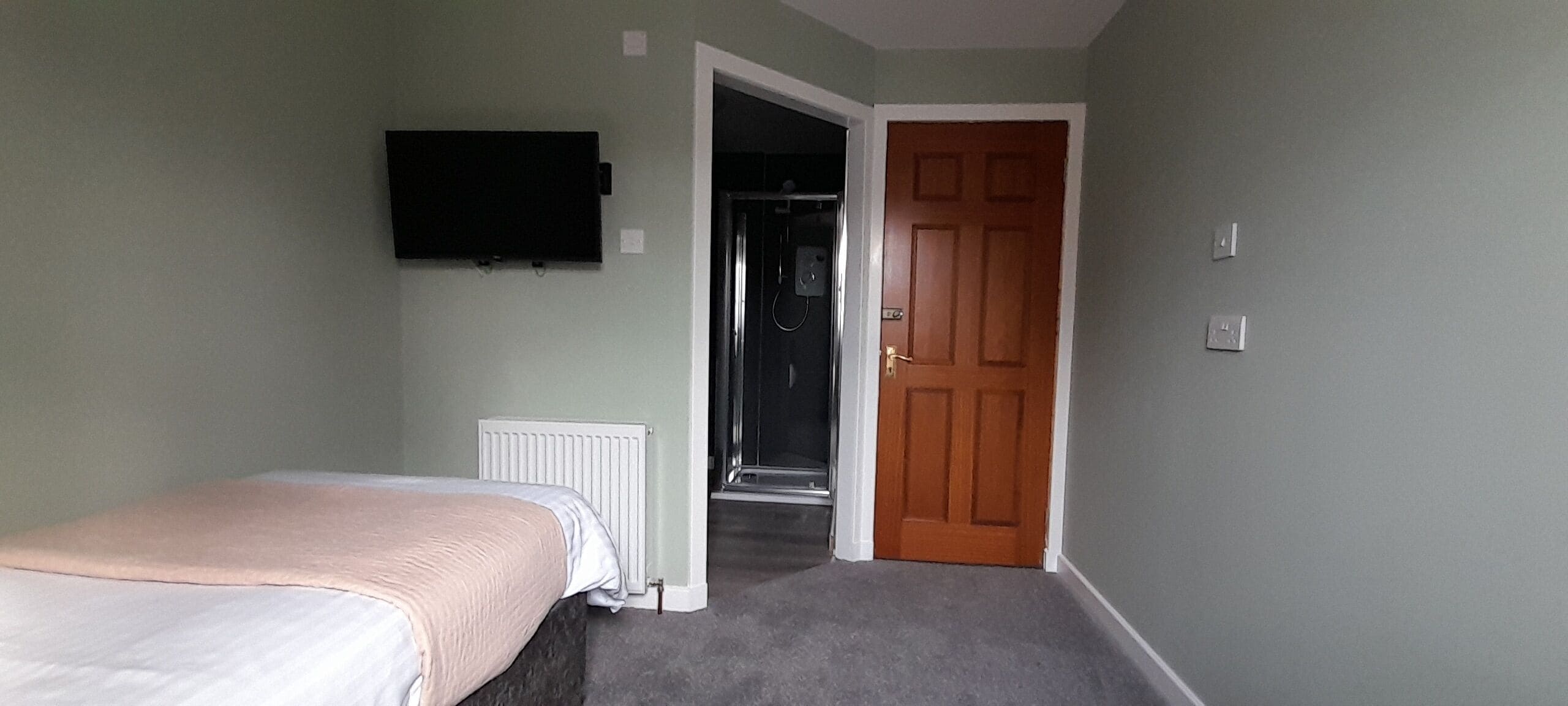 Un dormitorio con una cama, un televisor instalado en la pared y una puerta que da a un pasillo.