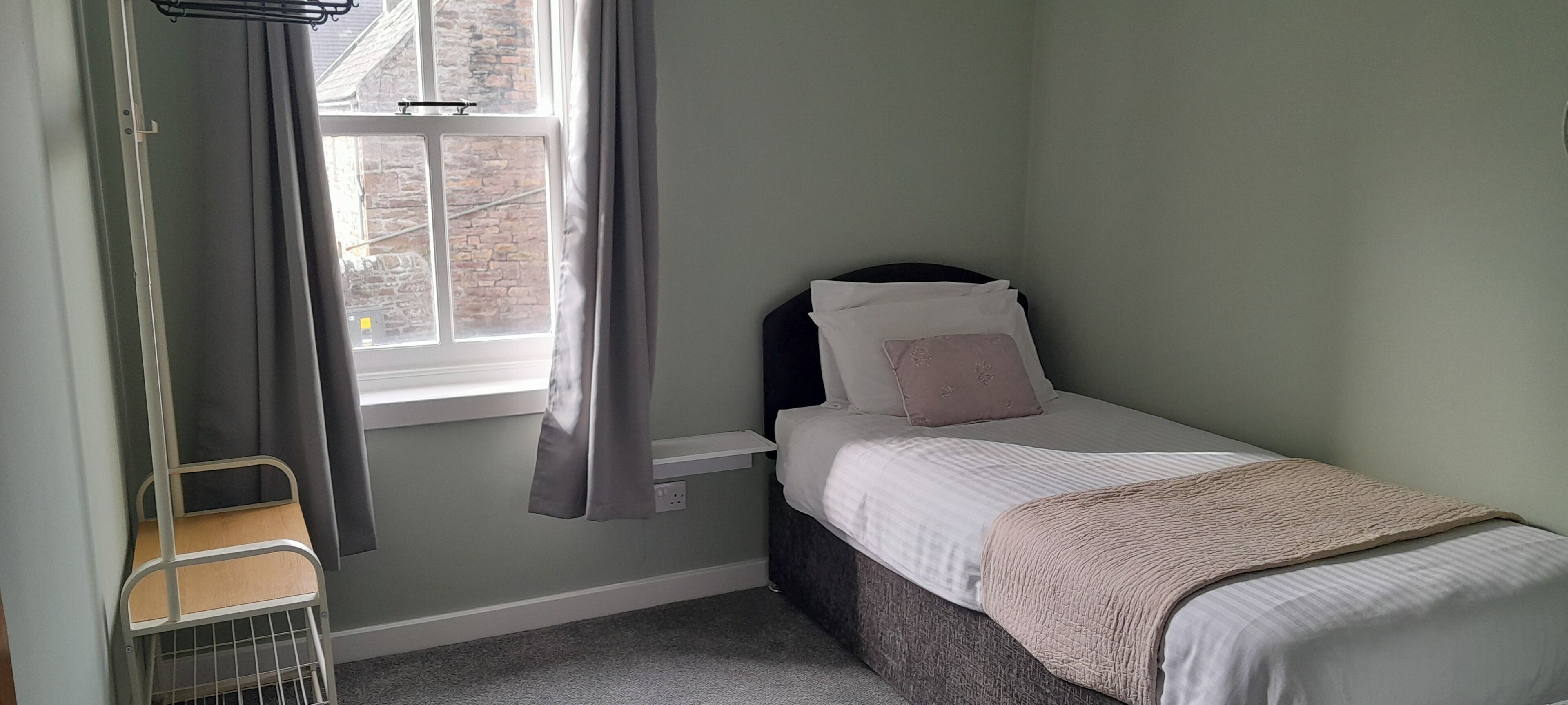 Un dormitorio pequeño y ordenado con una cama individual, una ventana con cortinas y una estantería metálica.